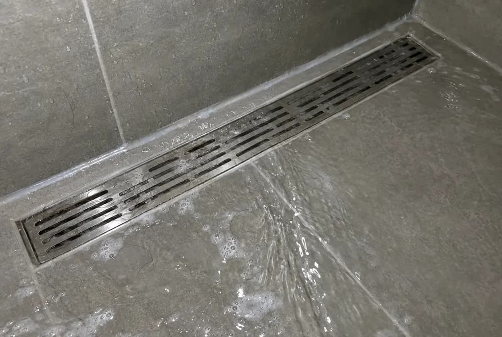 Sumidero lineal de acero inoxidable enrasado con el porcelánico gris de una ducha, con el agua corriendo hacia la rejilla