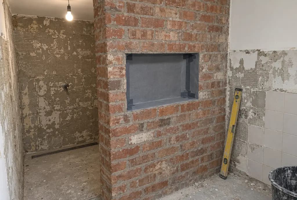 Hueco rectangular abierto en un tabique de ladrillo durante una obra de baño, con el interior cubierto por lámina impermeabilizante gris y esquinas selladas