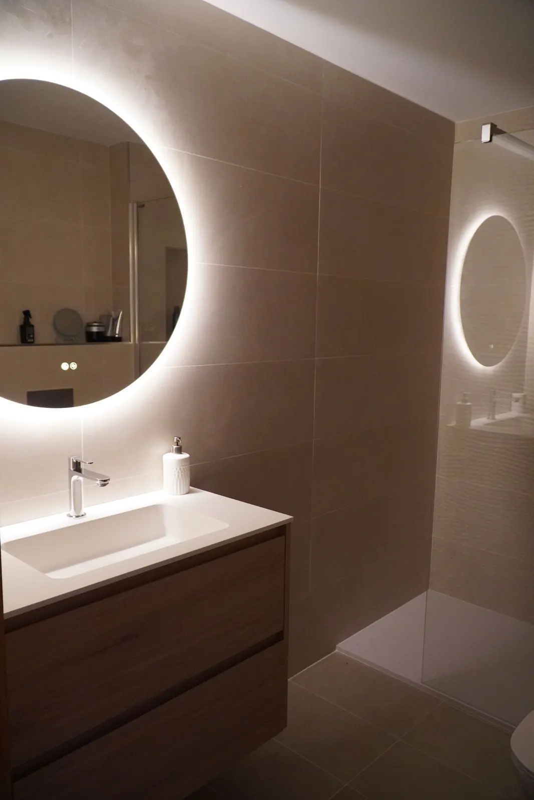 Baño reformado con espejo redondo retroiluminado y microcemento beige, resultado moderno